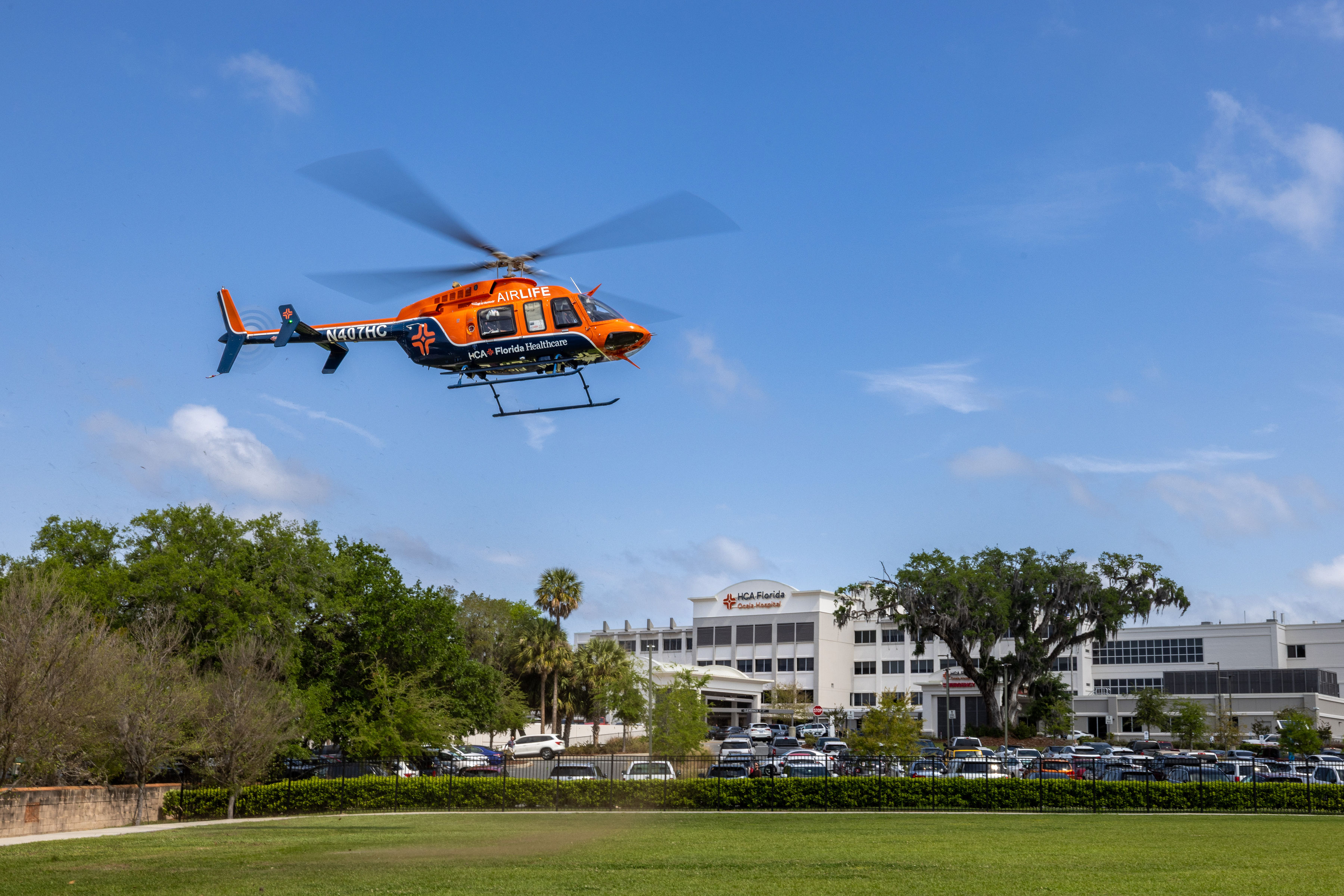 An orange AirLife medical helicopter flies above a hospital campus with a clear blue sky in the background.