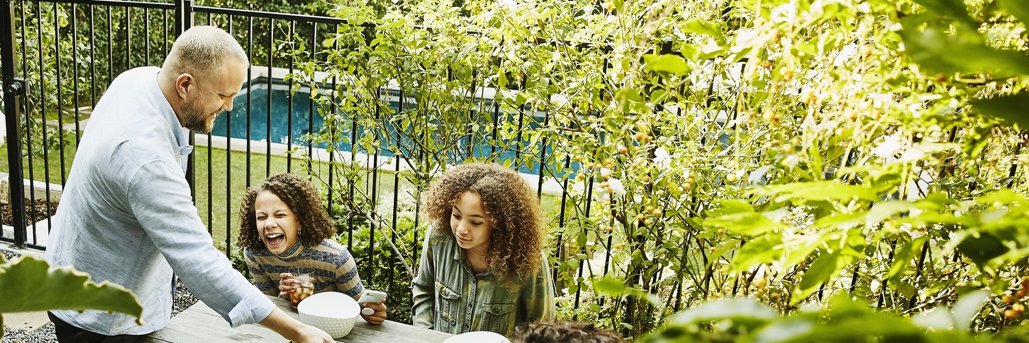 A smiling and laughing family of four shares a meal at a picnic table in a courtyard outside, with a swimming pool in the background.